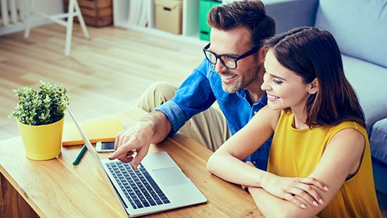Couple souriant consultant un site internet sur un ordinateur portable posé sur la table de leur salon