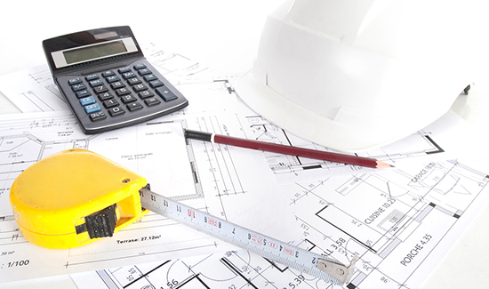Un casque de chantier, une calculatrice, un crayon et un mètre posés sur les plans d'un logement